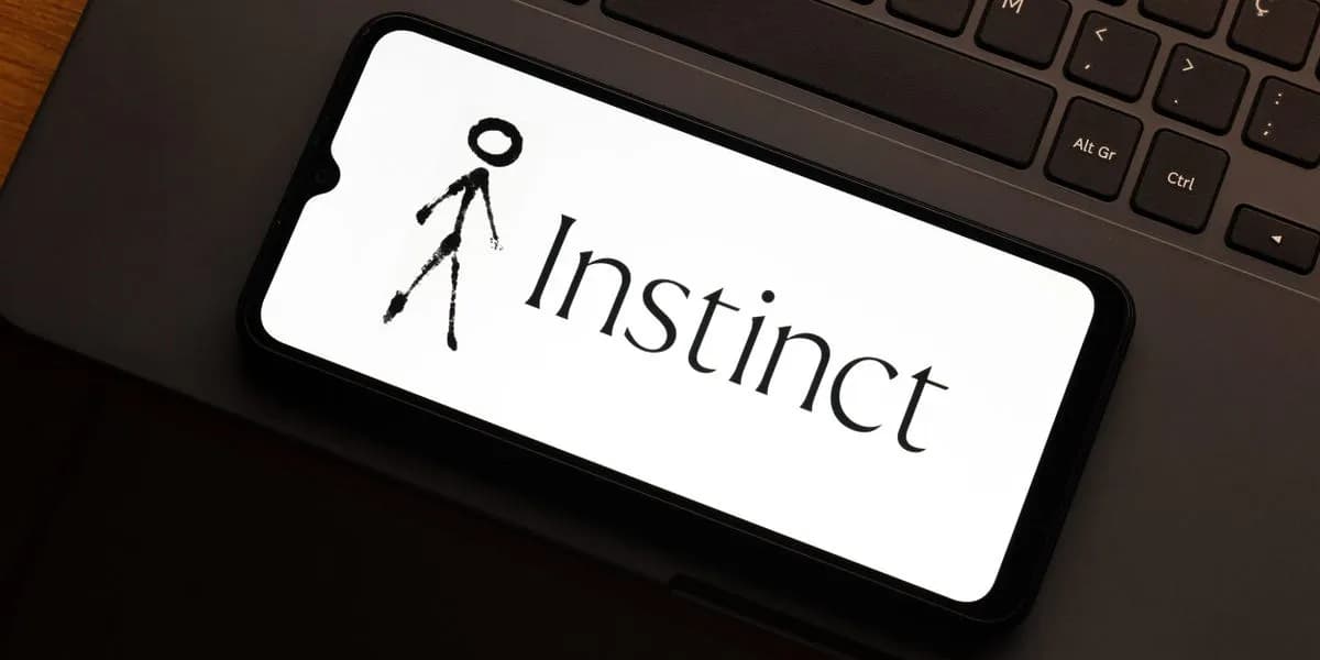 لوگوی Instinct، دستیار شخصی هوش مصنوعی، روی صفحهٔ یک گوشی هوشمند کنار کیبورد لپ‌تاپ
